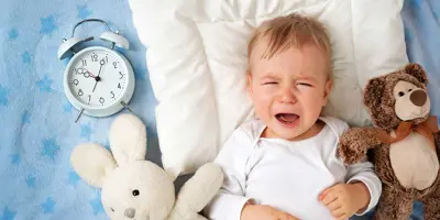 A baby crying on a blanket surrounded by two cuddly toys and an analogue alarm clock