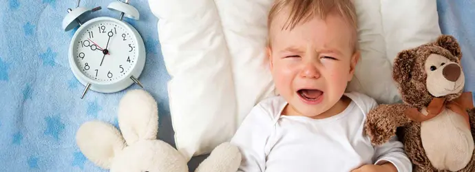 A baby crying on a blanket surrounded by two cuddly toys and an analogue alarm clock