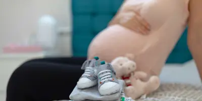 A neat pile of baby's clothes on a bed with a pregnant women sat in the background