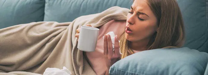 Woman lying in the sofa coughing while holding tea cup and a tissue