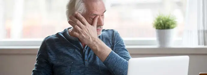 Elderly man in front of a laptop itching one eye