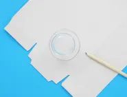 A tissue box opened flat is laid on a blue surface with an empty tumbler and pencil.