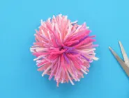 A pom pom made with different shades of pink wool, with a pair of scissors on the side.